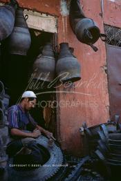 Image du Maroc Professionnelle de  Un Homme découpe un pneu de camion pour le transformer  en différents objets dont quelques uns sont accrochés au dessus. Plusieurs ateliers existaient dans rue situés derrière le cinéma Mabrouka où quelques artisans transformaient des pneus usagés en sceau pour hammam, cruche ou jarre que les paysans utilisaient pour faire sortir l’eau des puits. A cette époque le recyclage des pneus faisait partie intégrante du paysage social au Maroc, presque tous les hammams ont utilisés ces sceaux en caoutchouc à la place de ceux en bois, à cause de leur prix exorbitant et surtout qui se brisaient souvent par les lancements des ouvriers aux heures du nettoyage, Mercredi 18 Mai 1988. (Photo / Abdeljalil Bounhar)